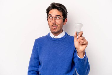 Beyaz arka planda izole edilmiş ampul tutan genç beyaz bir adam kafası karışmış, şüpheli ve güvensiz hissediyor..