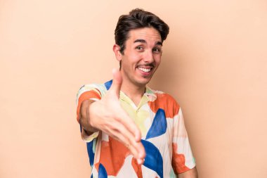 Beyazlı genç bir adam bej arka planda izole edilmiş. Kameraya doğru elini uzatarak selam veriyor..