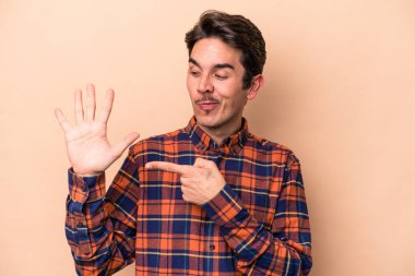 Beyaz tenli genç adam bej arka planda izole edilmiş. Gülümsüyor. Beş numarayı parmaklarıyla gösteriyor..