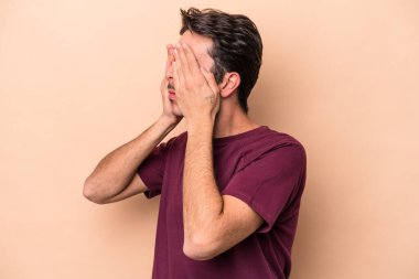 Beyazlı genç bir adam bej arka planda izole edilmiş, elleriyle gözlerini kapatmaktan korkuyor..