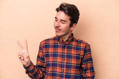 Beyaz tenli genç adam bej arka planda izole edilmiş neşeli ve kaygısız parmaklarıyla barış sembolü gösteriyor..