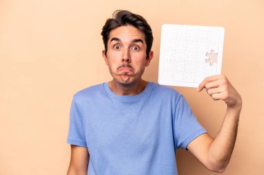 Kafkasyalı genç bir adam bej arka planda izole edilmiş bir yapboz tutuyor omuzlarını silkiyor ve gözleri açık..