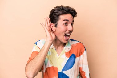 Beyazlı genç bir adam bej arka planda izole edilmiş bir şekilde bir dedikoduyu dinlemeye çalışıyor..