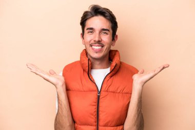 Beyazlı genç bir adam bej arka planda izole edilmiş, kollarıyla ölçek yapıyor, mutlu ve kendinden emin hissediyor..