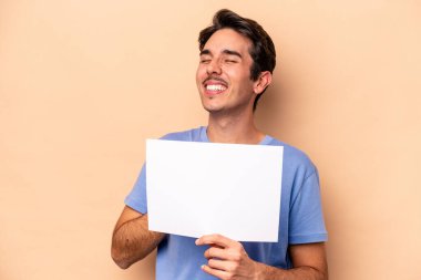 Beyazlı genç bir adam bej arka planda izole edilmiş bir pankart tutuyor ve elini göğsünde tutarak gülüyor..