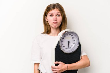 Beyaz arka planda izole edilmiş bir tartı tutan genç bir İngiliz kadın omuzlarını silker ve gözleri açık kalır..