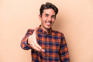 Beyazlı genç bir adam bej arka planda izole edilmiş. Kameraya doğru elini uzatarak selam veriyor..