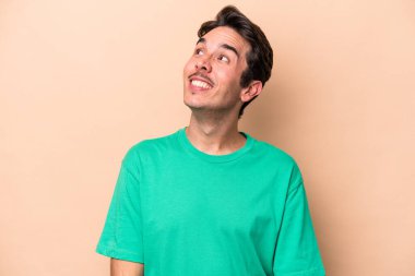 Beyaz tenli genç adam bej arka planda izole edilmiş rahat ve mutlu bir şekilde gülüyor, boynunda dişlerini gösteriyor..