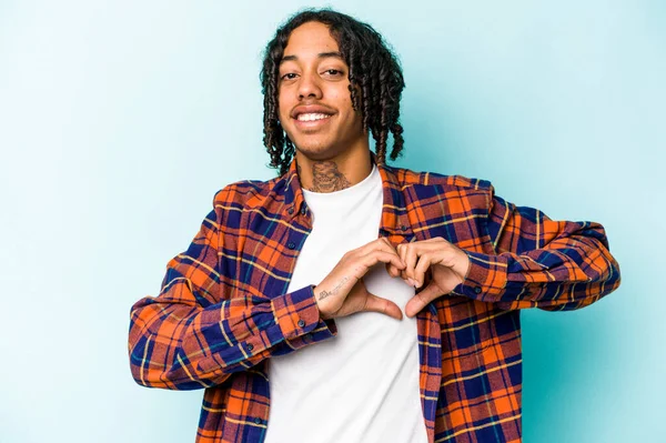 Genç Afrikalı Amerikalı adam mavi arka planda izole edilmiş gülümsüyor ve elleriyle kalp şekli gösteriyor..