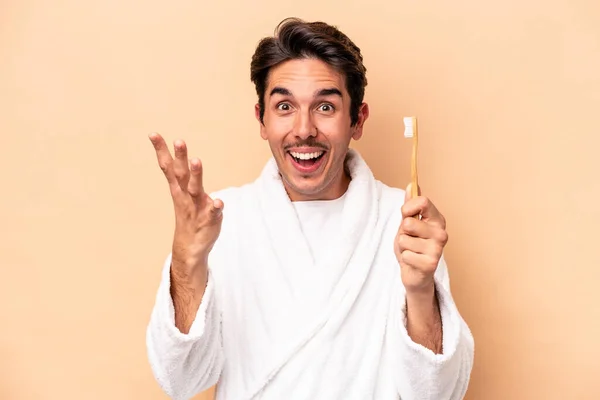 身穿浴衣的年轻的高加索人拿着一把用米色背景隔开的牙刷 带着惊喜 兴奋地举起双手 — 图库照片