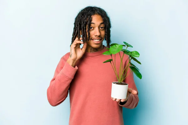 Young African American man holding a plant isolated on blue background trying to listening a gossip.