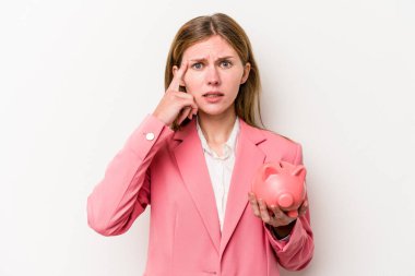 Beyaz arka planda izole edilmiş domuz kumbarası tutan genç İngiliz kadın işaret parmağıyla hayal kırıklığı jesti gösteriyor..