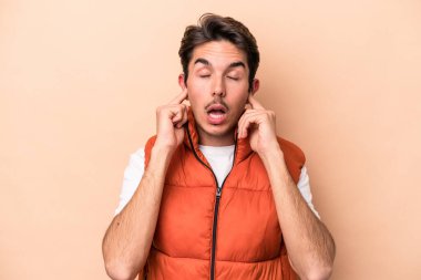 Beyazlı genç bir adam bej arka planda izole edilmiş parmaklarıyla kulaklarını kapatıyor, gürültülü bir ortam yüzünden stresli ve çaresiz..