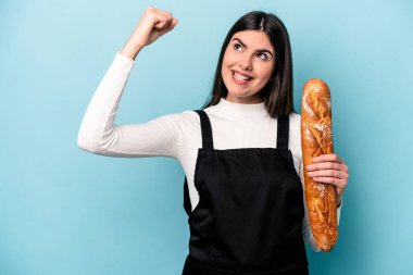 Genç fırıncı Kafkas elinde mavi arka planda izole edilmiş bir somun ekmek zaferden sonra yumruğunu kaldırıyor..