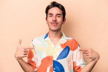Beyazlı genç bir adam bej arka planda izole edilmiş. Elle bir gömlek fotokopi alanını işaret ediyor. Gururlu ve kendinden emin.