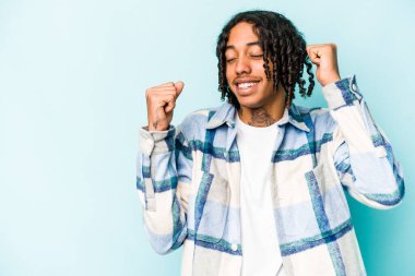 Genç Afrikalı Amerikalı bir adam mavi arka planda izole edilmiş zafer ve galibiyet kavramından sonra yumruğunu kaldırıyor..