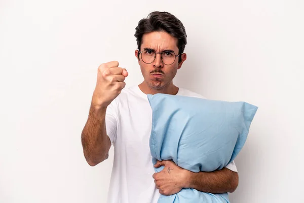 Young caucasian man wearing pajamas holding a pillow isolated on white background showing fist to camera, aggressive facial expression.