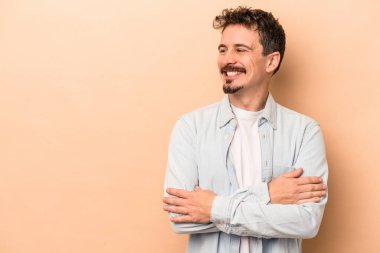 Beyaz tenli genç adam bej arka planda izole edilmiş. Kollarını kavuşturup gülümsüyor..