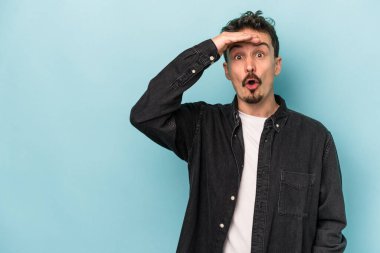 Kafkasyalı genç adam mavi arka planda izole edilmiş, uzaklara bakıyor ve elini alnına koyuyor..