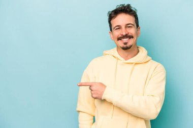 Mavi arka planda izole edilmiş genç beyaz adam gülümsüyor ve boş alanda bir şeyler gösteriyor..