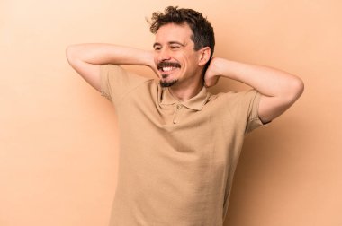 Beyazlı genç bir adam bej arka planda izole edilmiş. Kendine güveniyor, elleri başının arkasında..