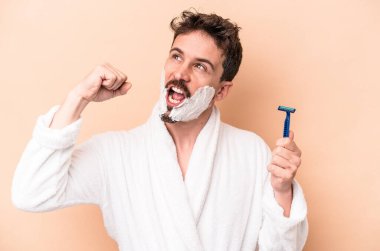 Tıraş köpüğü giymiş, elinde jilet tutan, bej arka planda saklanmış beyaz bir adam zafer kazandıktan sonra yumruğunu kaldırıyor..