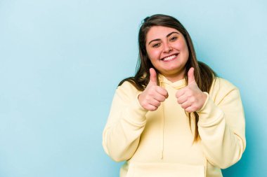 Beyaz, şişman bir kadın mavi arka planda izole edilmiş gülümsüyor ve başparmağını kaldırıyor.