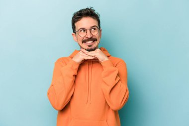 Mavi arka planda izole edilmiş genç beyaz adam ellerini çenesinin altında tutuyor, mutlu bir şekilde yana bakıyor..