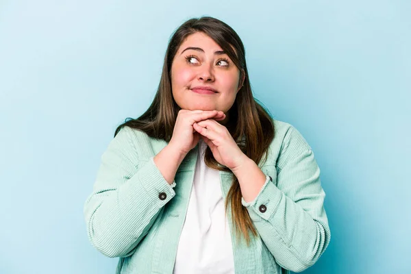 Young caucasian overweight woman isolated on blue background keeps hands under chin, is looking happily aside.