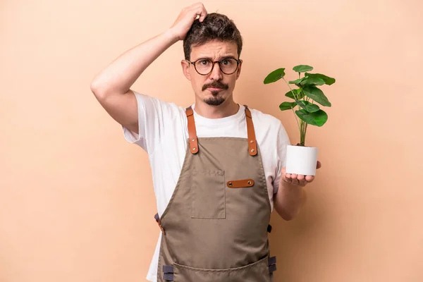 Young caucasian gardener man holding a plant isolated on beige background being shocked, she has remembered important meeting.