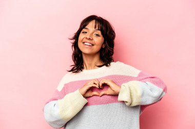 Pembe arka planda izole edilmiş genç İspanyol kadın gülümsüyor ve elleriyle kalp şeklini gösteriyor..