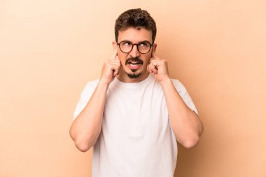 Beyazlı genç bir adam bej arka planda izole edilmiş parmaklarıyla kulaklarını kapatıyor, gürültülü bir ortam yüzünden stresli ve çaresiz..