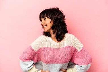 Pembe arka planda izole edilmiş genç İspanyol kadın mutlu bir şekilde gülüyor ve ellerini karnının üstünde tutarak eğleniyor..