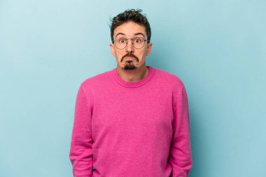 Mavi arka planda izole edilmiş genç beyaz adam omuzlarını silkeliyor ve gözleri açık..