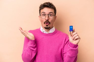 Elinde batarya tutan genç beyaz adam bej arka planda izole edilmiş. Şaşırmış ve şok olmuş..
