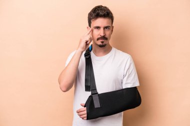 Kırık kollu genç beyaz adam bej arka planda izole edilmiş. Parmağıyla şakağı işaret ediyor. Düşünüyor, bir göreve odaklanıyor..
