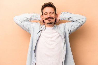 Beyazlı genç bir adam bej arka planda izole edilmiş. Kendine güveniyor, elleri başının arkasında..