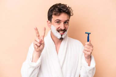 Tıraş köpüğü takan ve elinde jilet tutan beyaz bir adam bej arka planda iki numarayı parmaklarıyla gösteriyor..