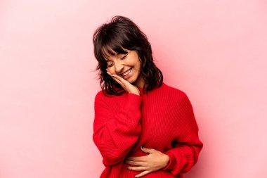 Pembe arka planda izole edilmiş genç İspanyol kadın mutlu bir şekilde gülüyor ve ellerini karnının üstünde tutarak eğleniyor..