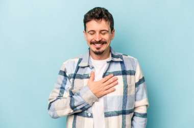 Mavi arka planda izole olmuş genç beyaz adam yüksek sesle gülüyor ve elini göğsüne koyuyor..