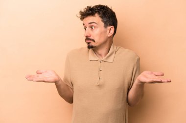 Kafkasyalı genç adam bej arka planda izole edilmiş. Kafası karışmış ve omuz silkiyor. Kopya alanını tutmak için..