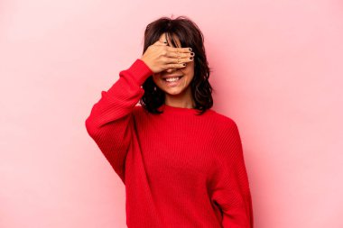 Pembe arka planda izole edilmiş genç İspanyol kadın elleriyle gözlerini kapatıyor, geniş bir gülümsemeyle bir sürpriz bekliyor..