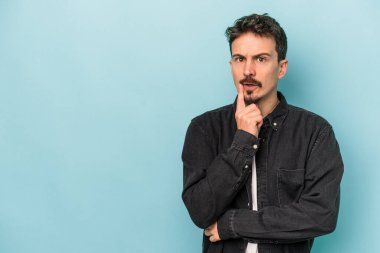 Mavi arka planda izole edilmiş genç beyaz adam şüpheli ve şüpheci bir ifadeyle yana bakıyor..