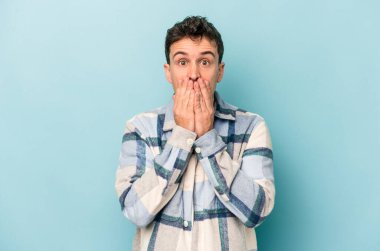 Mavi arka planda izole edilmiş genç beyaz adam şok olmuş, ağzı ellerle kaplanmış, yeni bir şey keşfetmeye hevesli..