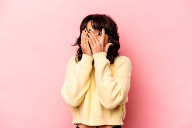 Pembe arka planda izole edilmiş genç İspanyol kadın parmaklarının arasından korkmuş ve gergin bir şekilde göz kırpıyor..