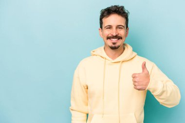 Mavi arka planda izole edilmiş genç beyaz adam gülümsüyor ve başparmağını kaldırıyor.
