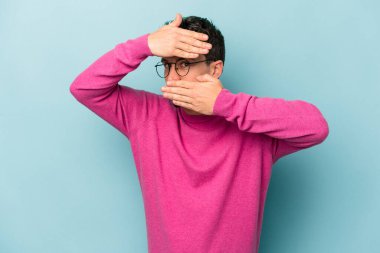 Mavi arka planda izole edilmiş genç beyaz adam kameraya parmaklarıyla göz kırpıyor, yüzünü kapatmaktan utanıyor..