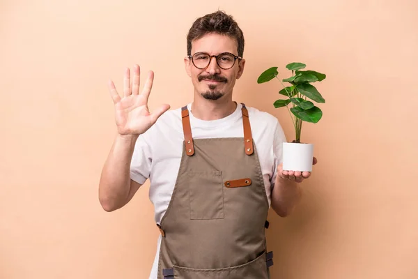 Young caucasian gardener man holding a plant isolated on beige background smiling cheerful showing number five with fingers.