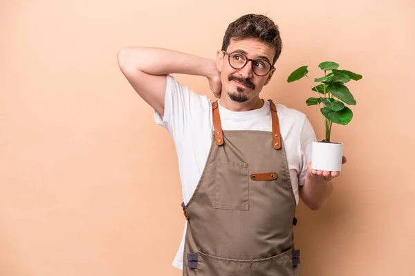 Young caucasian gardener man holding a plant isolated on beige background touching back of head, thinking and making a choice.