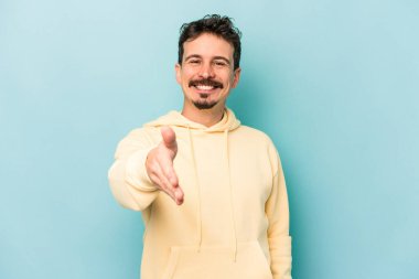 Mavi arka planda izole edilmiş genç beyaz adam selamlama jesti olarak kameraya elini uzatıyor..